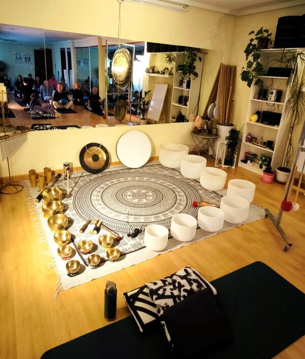 Salón con suelo de madera y colores cálidos. Sobre una alfombra con mandala se reparten cuencos tibetanos, cuencos de cuarzo, gongs y más instrumentos para sonoterapia. En un gran espejo en la pared se reflejan personas realizando ejercicios de relajación.